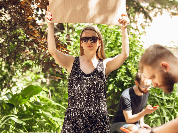 Frau mit unbeschriebenem Protestplakat, rundherum in Park starren junge Menschen auf ihre Smartphones