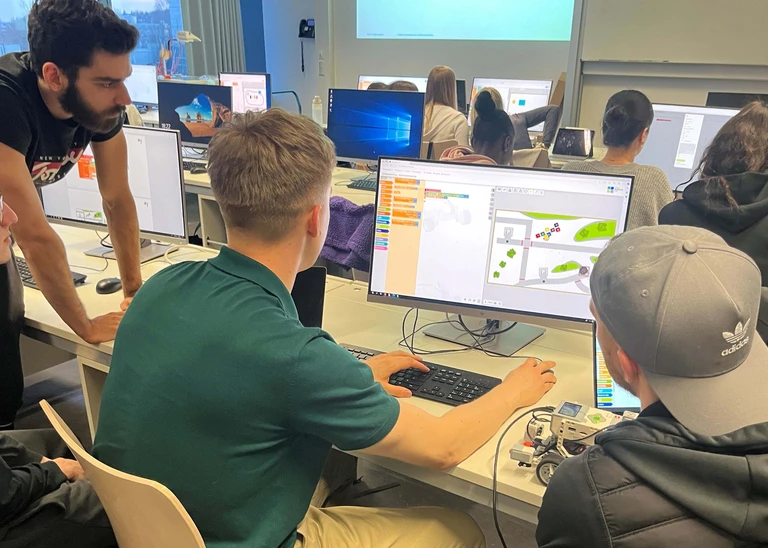 Mehrere Studierende sitzen um einen Computer und arbeiten zusammen an einer Programmierung.