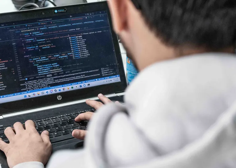 Man sieht über die Schulter eines Mannes auf seinen Laptop, wo er etwas programmiert. 