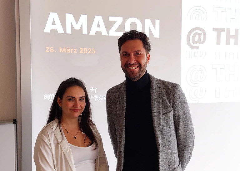 Eine Frau und ein Mann lächeln in die Kamera, hinter Ihnen sieht man eine Power Point Präsentation.