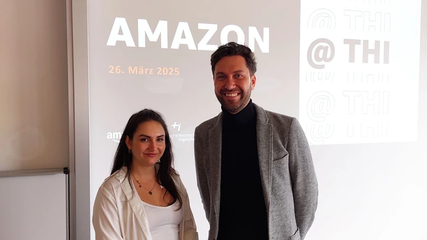 Eine Frau und ein Mann lächeln in die Kamera, hinter Ihnen sieht man eine Power Point Präsentation.