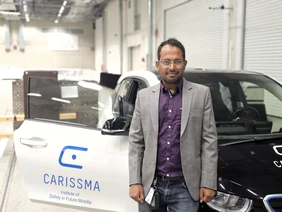 For more vehicle safety: Gerald Joy Sequeiras uses the CARISSMA hall for his driving tests (Photo: THI). Portrait mit Mann im Vordergrund, hinter Ihm ein Auto mit "CARISSMA"-Label auf der offenen Beifahrertüre