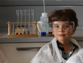 Like a real researcher: 5-year-old Vincent in the lab (Photo: THI). KiGa-Kind in einem Labor mit Schutzbrille und -Kleidung, im Hintergrund Reagenzgläser