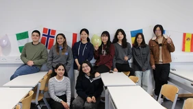 9 people stand in a classroom and smile into the camera, in the background you can see many different flags.