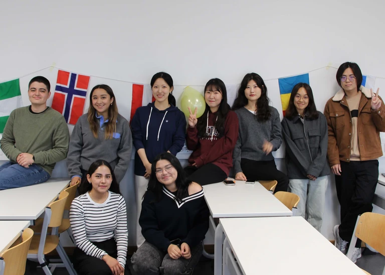 9 people stand in a classroom and smile into the camera, in the background you can see many different flags.