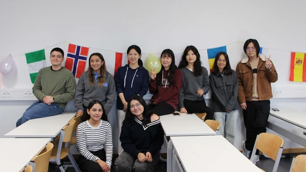9 people stand in a classroom and smile into the camera, in the background you can see many different flags.